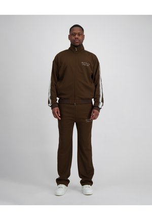 Man staat en draagt een bruin trainingspak met witte strepen op mouwen en pijpen, gecombineerd met witte sneakers, tegen een effen achtergrond.