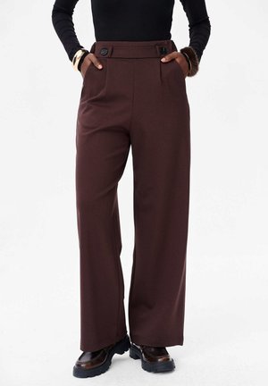 Persoon in hoge taille, wijdvallende, bruine broek met knoopdetails, zwarte langemouwen top en zwarte schoenen met dikke zool, met de handen in de zakken.