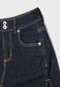 Jeans en denim bleu foncé avec une taille haute, deux boutons en métal et des coutures jaunes contrastantes. Poches standards à l'avant.