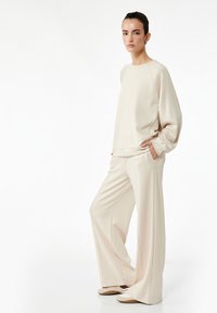 Sudadera de color beige claro con mangas raglán y bolsillos laterales, combinada con pantalones de pierna ancha a juego, que muestran una tela suave y texturizada.