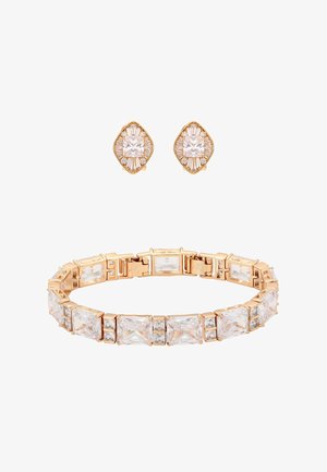 Bransoletka w złotym odcieniu z prostokątnymi i kwadratowymi przezroczystymi kryształami, charakteryzująca się elastycznym designem. Do kompletu pasujące kolczyki wkrętki z podobnymi kamieniami.