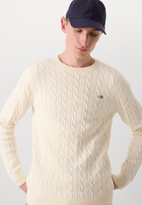 Maglione crema a trecce con collo rotondo e polsini a coste, caratterizzato da intricati motivi a treccia. Abbinato a un berretto da baseball blu navy.