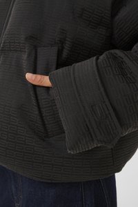 Svart puffjakke med en teksturert geometrisk mønster, med glidelås foran og hånden stukket ned i en sidelomme, med brede, brettede mansjetter.