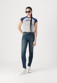 Camiseta gris corta con mangas azules y logo rojo, combinada con jeans ajustados azules y zapatillas blancas. Presenta gafas de sol y cabello trenzado.