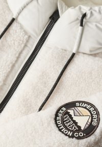 Veste en polaire blanche avec une fermeture éclair noire, une capuche ajustable et un écusson rond avec des graphiques de montagne et le texte "SUPERDRY EXPEDITION CO."