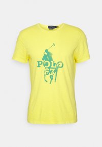 Keltainen puuvillapaita, jossa on turkoosi poloilija- grafiikka ja "POLO 67" -teksti. Klassinen pyöreä kaula-aukko ja lyhyet hihat. Pehmeä rakenne.