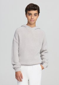 Jasnoszara dziana bluza z szerokim dekoltem, prążkowanymi mankietami i luźnym krojem. W połączeniu z białymi spodniami i neutralnym tłem.