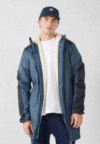 Giacca imbottita blu con maniche nere, foderata in pile crema. Presenta una chiusura con cerniera e un cappuccio, indossata sopra una camicia bianca e pantaloni scuri.