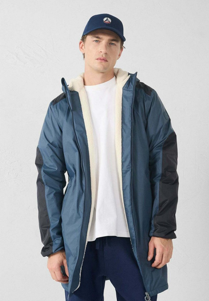 Giacca imbottita blu con maniche nere, foderata in pile crema. Presenta una chiusura con cerniera e un cappuccio, indossata sopra una camicia bianca e pantaloni scuri.