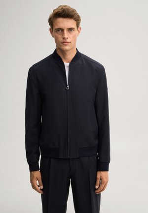 Giovane uomo con capelli castano chiaro indossa una giacca bomber nera con cerniera e pantaloni neri abbinati su uno sfondo chiaro e semplice.