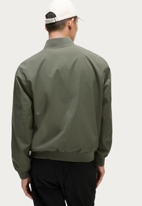 Chaqueta bomber verde oliva con cuello y puños acanalados. Textura lisa y mate, con una cintura ajustada y un diseño minimalista.
