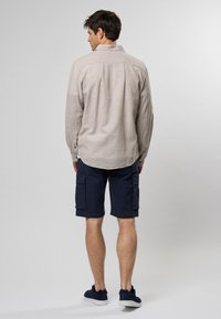 Camisa de manga larga color beige claro con un acabado texturizado, combinada con pantalones cortos cargo azul marino con bolsillos laterales. El modelo lleva zapatillas de ante azul marino.
