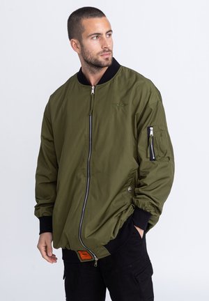 Olivgrüne Bomberjacke mit einem Frontreißverschluss, Rippbündchen und einem elastischen Saum. Verfügt über eine Utility-Tasche am linken Ärmel und ein orangefarbenes Etikett.