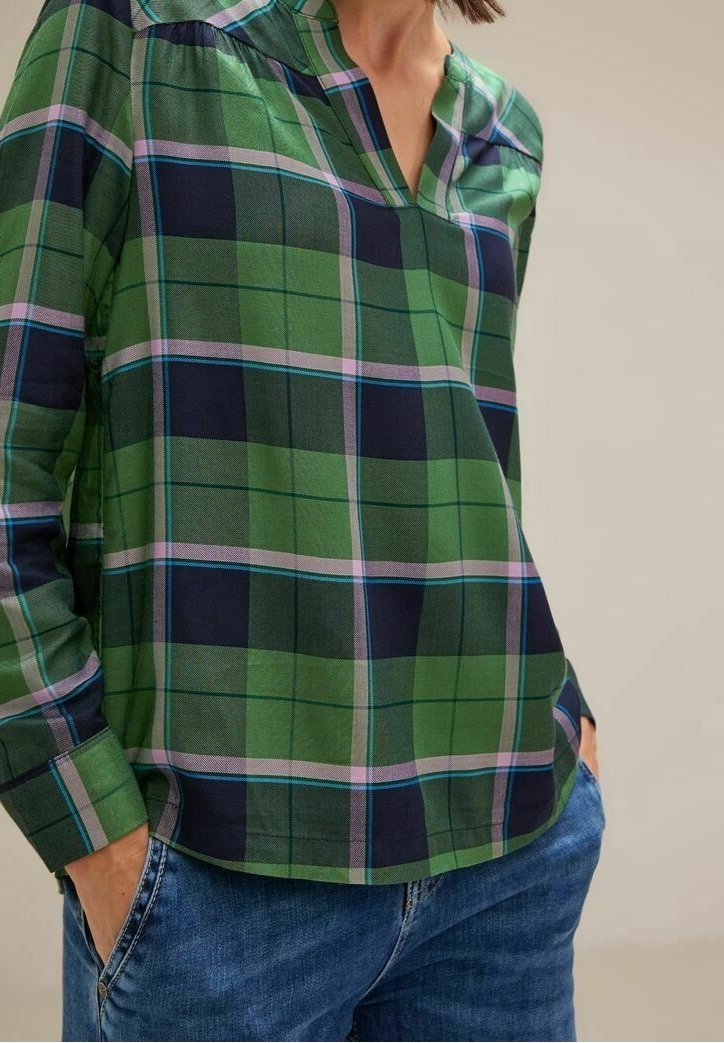 Femme portant une chemise à carreaux verte, bleue et violette à manches longues avec un col en V, les mains dans les poches, associée à un jean bleu.
