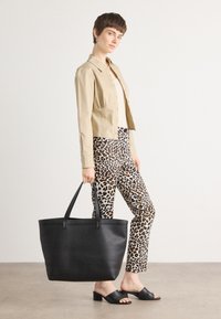 Sac fourre-tout en cuir noir avec deux poignées, texture lisse et forme structurée. Le modèle porte une veste beige courte et un pantalon à imprimé animal.