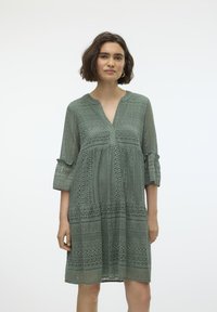Robe en dentelle verte avec un décolleté en V, des manches trois-quarts et des poignets volantés. Présente des motifs intriqués et une coupe décontractée. Texture légère et aérée.