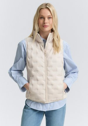 Blondynka ubrana w jasnobeżową pikowaną kamizelkę na niebiesko-białej pasiastym koszuli zapinanej na guziki i jasnoniebieskich dżinsach, stojąca z rękami w kieszeniach.