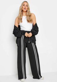 Zwarte wijde pantalons met witte zijstrepen, gecombineerd met een aansluitend wit tanktop en een zwarte bomberjack met ribbels bij de manchetten en kraag.