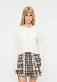 Κρεμ ζακέτα με V-λαιμόκοψη, υφασμένο ανάγλυφο πλέξιμο, μπροστινό κούμπωμα με ένα κουμπί, σε συνδυασμό με καφέ καρό πλισέ φούστα που παρουσιάζει διασταυρούμενες γραμμές.