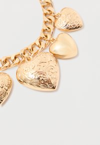 Ogrlica od zlatnog tona sa lancem koja sadrži tri privjeska u obliku srca različitih veličina, dva s kompleksnim gravurama i jedan s glatkom površinom.