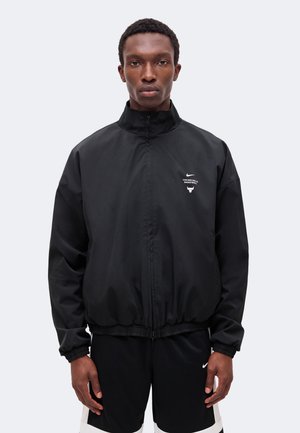 Un homme porte une veste de basket noire Nike Chicago Bulls zippée et un short assorti, sur un fond clair et uni.