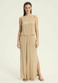 Beżowy kombinezon bez rękawów o swobodnym kroju, z prostym dekoltem, bocznymi rozcięciami i luźnym fasonem. Lekka tkanina.