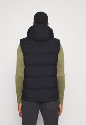 Man draagt een zwart gewatteerd hooded vest over een olijfgroen shirt met lange mouwen, staand voor een effen lichte achtergrond.