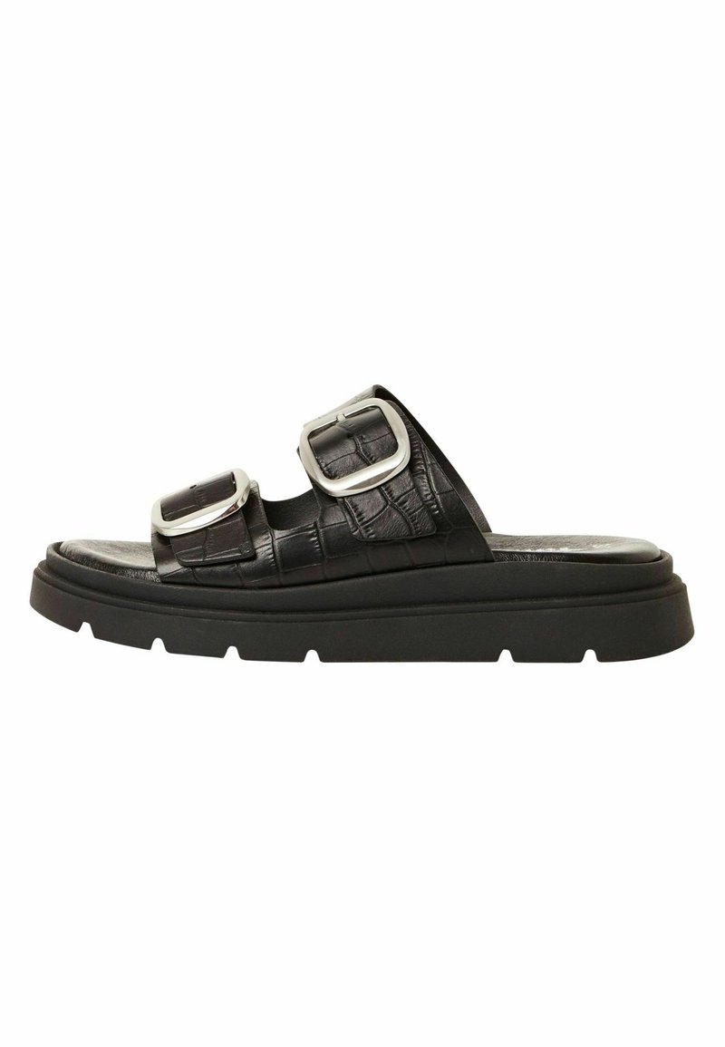 Sandale noire à semelle épaisse, avec deux larges brides texturées et de grandes boucles en argent sur fond blanc.