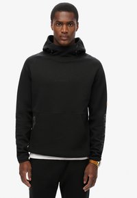Sudadera negra con un alto cuello, cordones, puños acanalados y un bolsillo tipo canguro en la parte frontal. Presenta un logo texturizado en el pecho y detalles en las mangas.