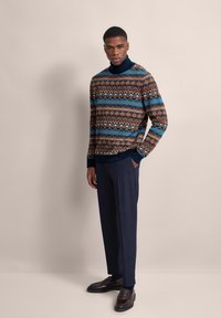 Brauner und blauer gemusterter Pullover mit hohem Kragen, kombiniert mit marineblauen Hosen und dunklen Slippern. Glatte Textur, geometrische Designs und kontrastierende Bündchen.