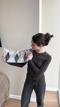 Vrouw met krullend haar, gekleed in een donker shirt met lange mouwen en een spijkerbroek, die een verpakte deken met ruitpatroon binnenshuis vasthoudt.