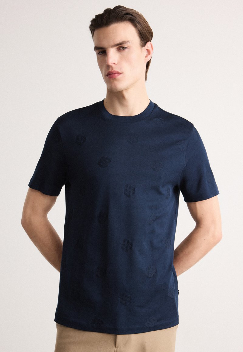 Tamsiai mėlyna trumpomis rankovėmis marškinėliai iš minkštos medžiagos, su subtiliu raštu ir apvalia iškirpte. Baigimas apima tiesų apatinį kraštą.