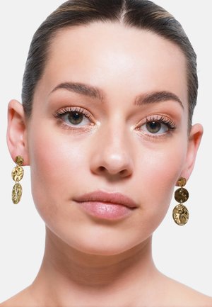 Goldene Metall-Ohrringe mit einem strukturierten Design, das aus drei miteinander verbundenen kreisförmigen Formen besteht. Die Ohrringe hängen unterhalb des Ohrläppchens.