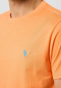 Tricou cu mânecă scurtă din bumbac portocaliu strălucitor, având un logo brodat albastru pe piept. Design minimalist cu guler rotund.