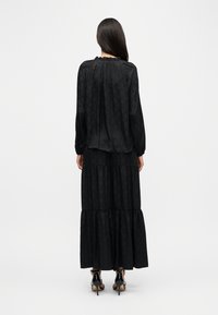 Μαύρο μακρύ φόρεμα από υφασμάτινη ύφανση με στρωμένη σχεδίαση, μακριά μανίκια και λαιμόκοψη με βολάν. Φορεμένο με παπούτσια με ψηλό τακούνι.