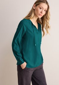 Blusa leggera color teal con scollo a V e cordoncino, caratterizzata da maniche lunghe con polsini elasticizzati. Abbinata a pantaloni grigio scuro.