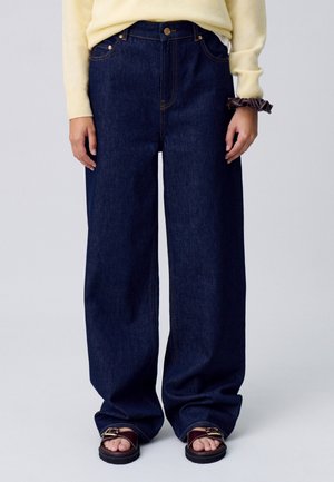 Jeans de denim de pierna ancha en azul oscuro con costuras naranjas en contraste, que cuentan con un cierre de botón y un diseño clásico de cinco bolsillos.
