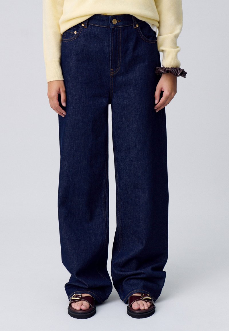 Jeans de denim de pierna ancha en azul oscuro con costuras naranjas en contraste, que cuentan con un cierre de botón y un diseño clásico de cinco bolsillos.