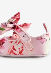 Zapato para bebé con patrón floral en tonos rosas, hecho de tela suave, que presenta un gran lazo en la parte superior, una correa ajustable y una suela texturizada.
