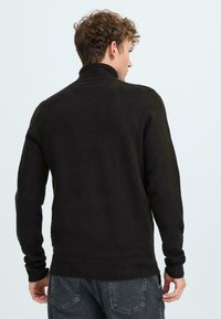 Črn pulover s visokim ovratnikom, izdelan iz teksturirane pletene tkanine. Ima dolge rokave in nekoliko ohlapen kroj, prikazan zadaj.