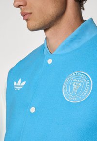 Light blue fleece varsity jacket with white accents, featuring embroidered Miami football club logo and Adidas three-stripe logo on the chest.