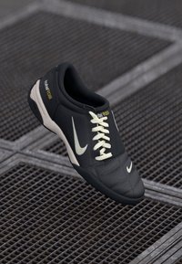 Crna i bijela nogometna kopačka Nike Total 90 III s bijelim vezicama, smještena iznad metalne rešetkaste površine.