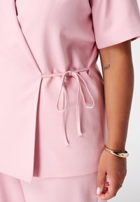 Lichtroze wikkeltop van textiel met structuur, met een strik in de taille. Korte mouwen en een loose fit. Gouden armband zichtbaar op de arm.
