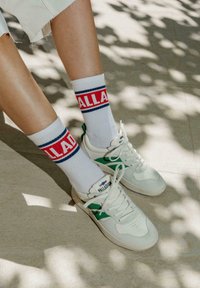 Sneakers brancos com accents verdes, apresentando um cabedal texturizado em suede e pele, combinados com meias brancas com riscas vermelhas e azuis e texto.