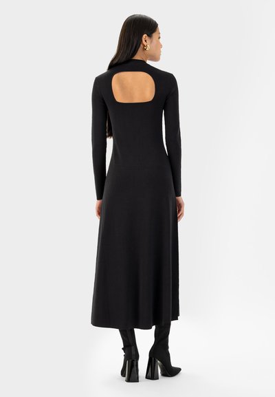 Robe noire à manches longues de longueur midi avec un col haut et un dos ouvert. Texture lisse et silhouette ajustée, associée à des bottes montantes.
