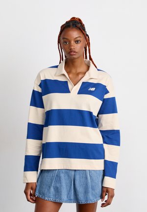 Woman with long orange braids wearing a blue and white striped long-sleeve rugby shirt and a pleated blue denim mini skirt, standing against a white background.