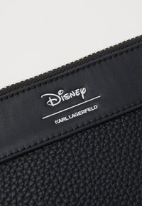 Črna usnjena torbica z teksturo in srebrnim zadrgo. Na sprednji strani je logotip: "Disney" v belem pisanju in "KARL LAGERFELD" v krepkem sans-serif napisu.