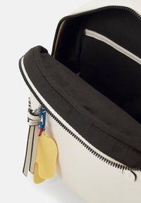 Krämmfärgad läderväska med svart foder, försedd med dragkedjestängning, innerv pocket och en gul charm fäst vid dragkedjan.