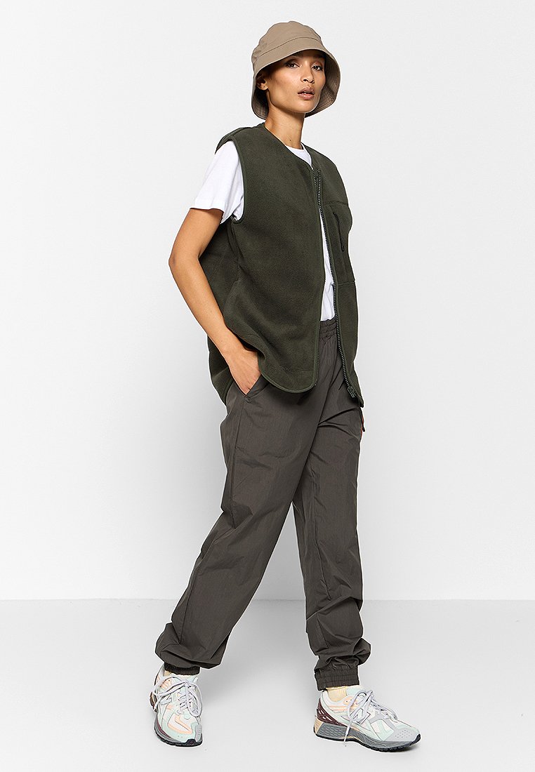 Olivgrüner Fleece-Weste mit einem Reißverschluss vorne, kombiniert mit grauen Hosen und mehrfarbigen Turnschuhen. Lässiger, lockerer Schnitt. Bucket Hat als Accessoire.