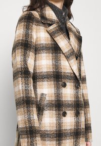 Femme portant un manteau à carreaux croisé avec de larges revers beige, noir et blanc par-dessus un pull zippé sombre.
